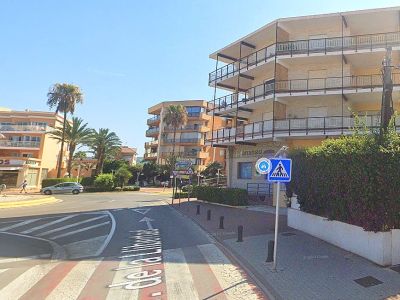 Edificio residencial en esquina con fachada clara y múltiples balcones, situado junto a un cruce de calles con paso de peatones y vegetación.
