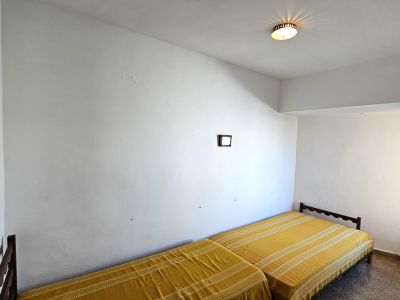Habitación con dos camas individuales y cabeceros de madera. Las paredes blancas muestran signos de uso, con una ventana lateral que aporta luz natural y un suelo de terrazo.