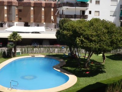 Piscina comunitaria de forma ovalada con bordillo claro, rodeada de césped verde y árboles que ofrecen sombra natural. Al fondo, aparcamiento cubierto y edificios residenciales.