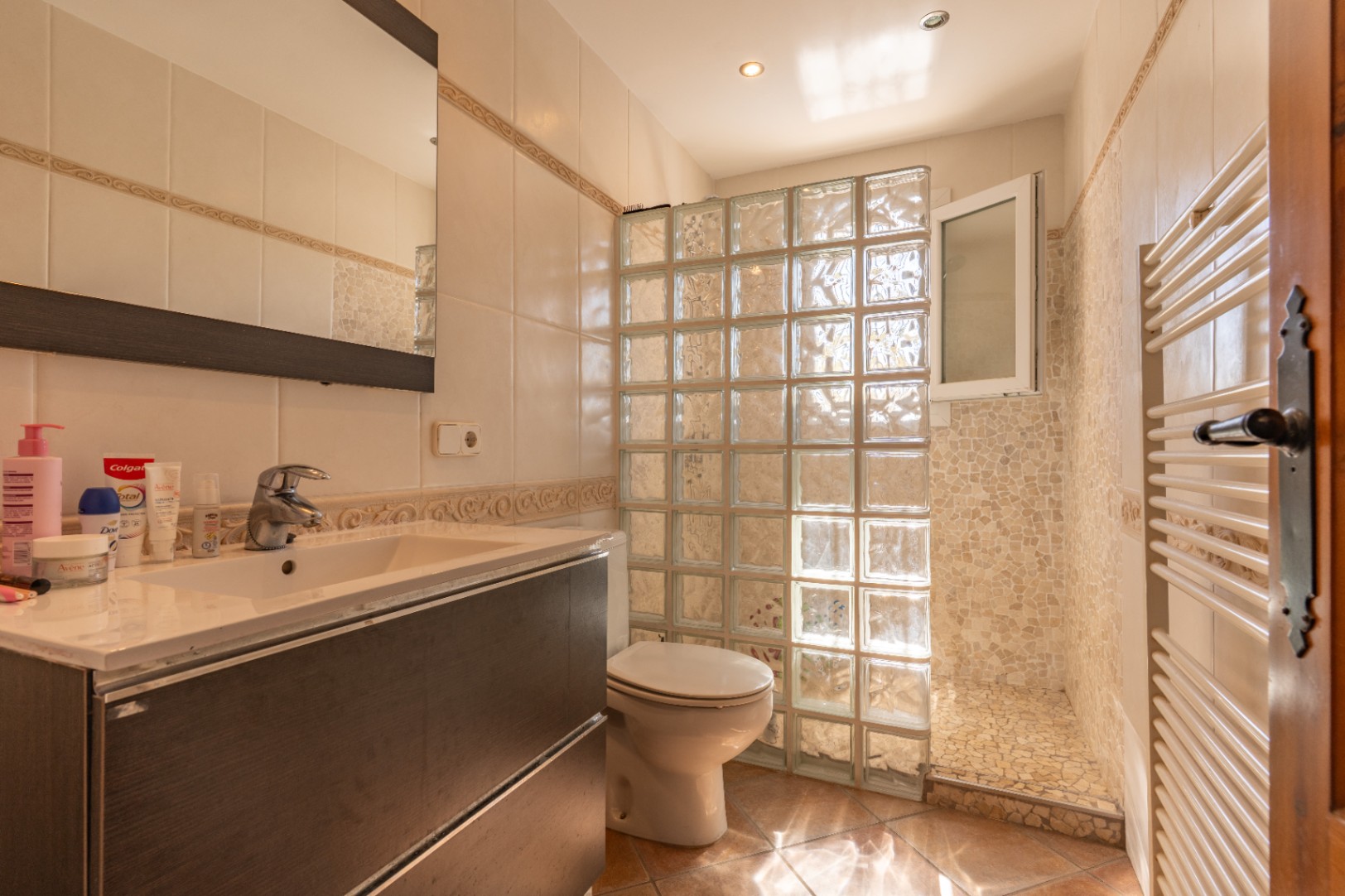Baño funcional con mueble de lavabo oscuro, mampara de bloques de vidrio para la ducha y zona de ducha con azulejos de mosaico. Incluye toallero calefactado y suelo de terracota.