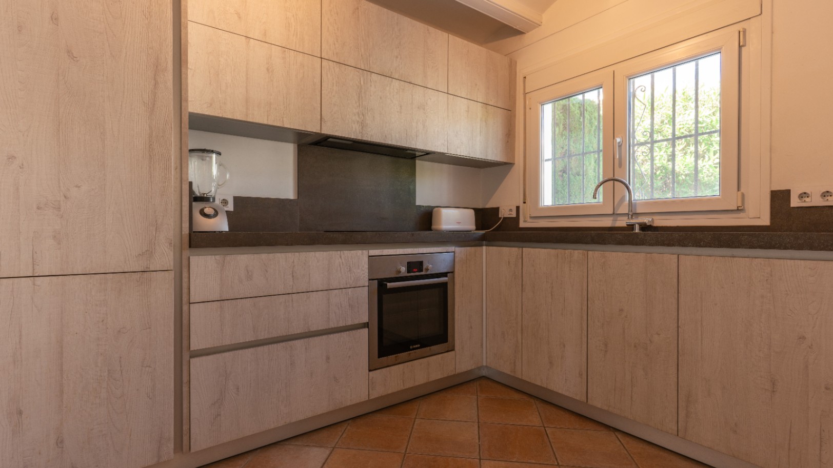 Cocina renovada en L con armarios efecto madera clara, encimeras grises oscuras y horno integrado. Ventana de PVC blanco con rejas aporta luz natural.