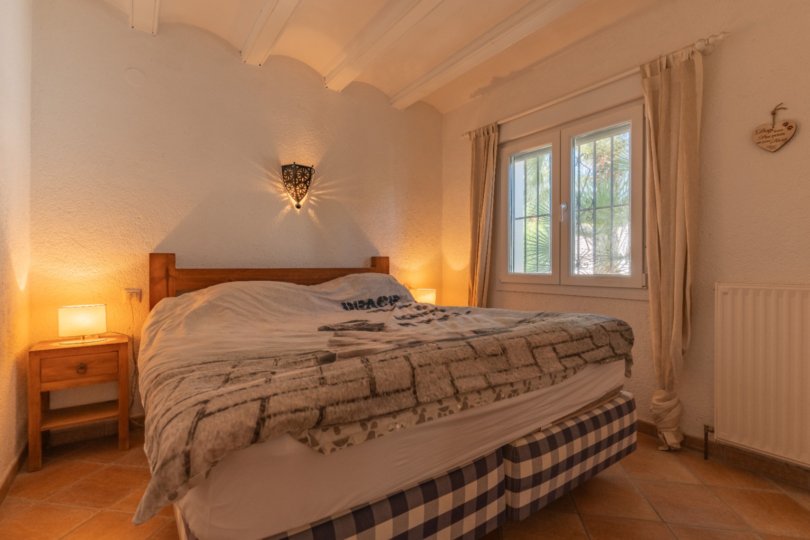 Dormitorio con cama de madera y base a cuadros, suelos de terracota y ventana de PVC con doble cristal. Destaca el aplique de pared con luz decorativa y el techo con vigas vistas.