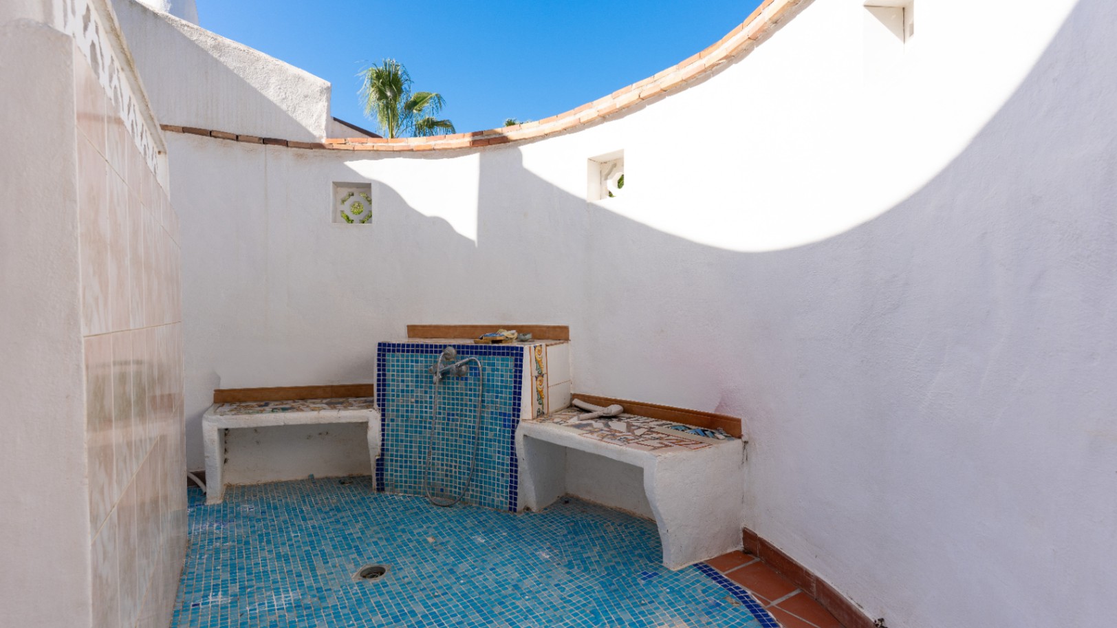 Ducha exterior con paredes blancas enlucidas y suelo de mosaico azul. Incluye bancos integrados con azulejos decorativos y una pared curva bajo el cielo azul.