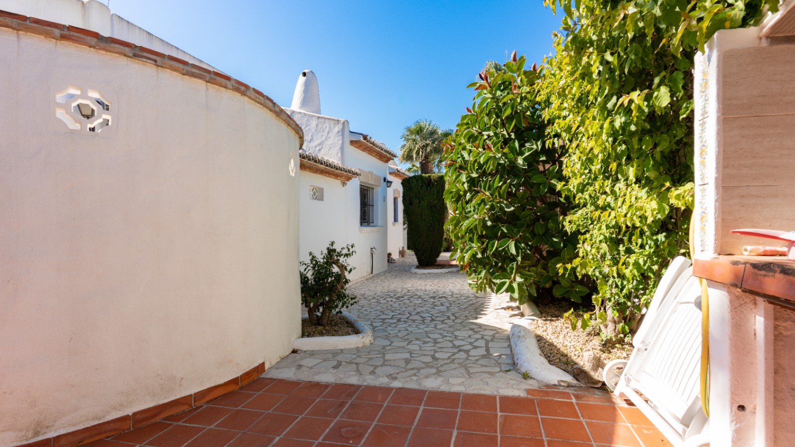 Entrada a la propiedad con un camino de piedra irregular, flanqueado por una pared curva blanca con celosía y exuberante vegetación mediterránea. Suelos de terracota en primer plano.