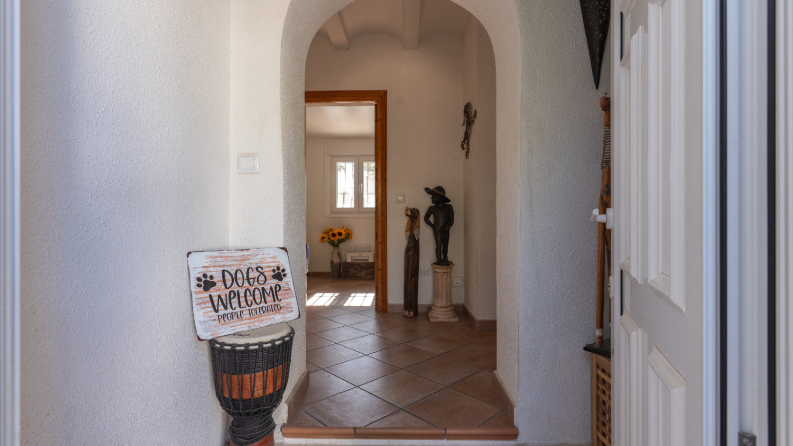 Entrada principal con arcos enlucidos en blanco y suelo de baldosas de terracota. La puerta de entrada panelada se ve a la derecha, y al fondo se divisa una estancia con luz natural y girasoles.