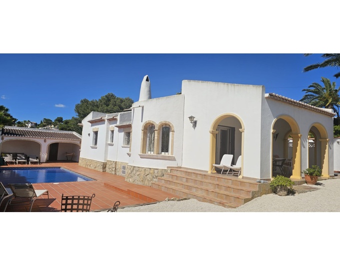 Exterior de villa blanca con arquitectura mediterránea, destacando una terraza cubierta con arcos ocres y escaleras de terracota. Piscina azul y zona de aparcamiento lateral.