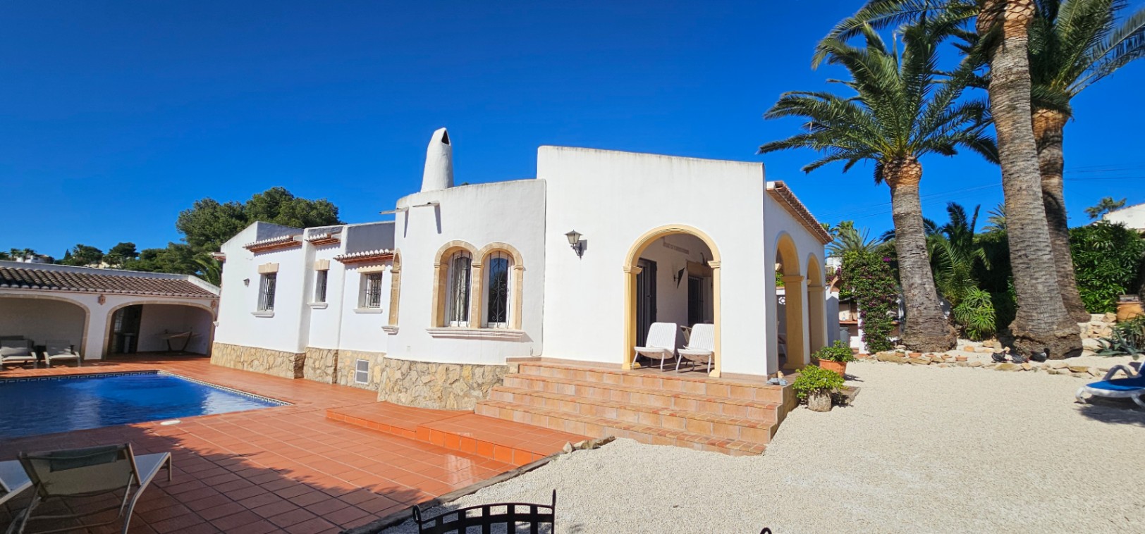 Exterior de villa blanca con base de piedra, escalones de terracota y porche arqueado. Piscina con terraza de gres y jardín mediterráneo con palmeras bajo cielo azul.