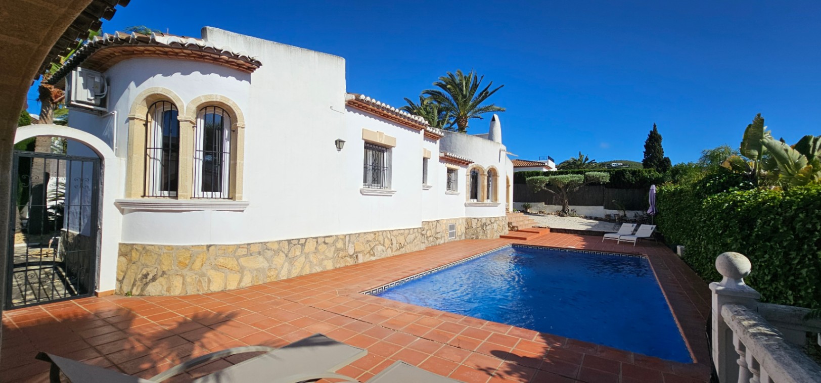 Exterior de villa blanca con base de piedra y ventanas arqueadas con rejas. Piscina rectangular rodeada de terraza de barro cocido, palmeras y vegetación bajo un cielo azul.