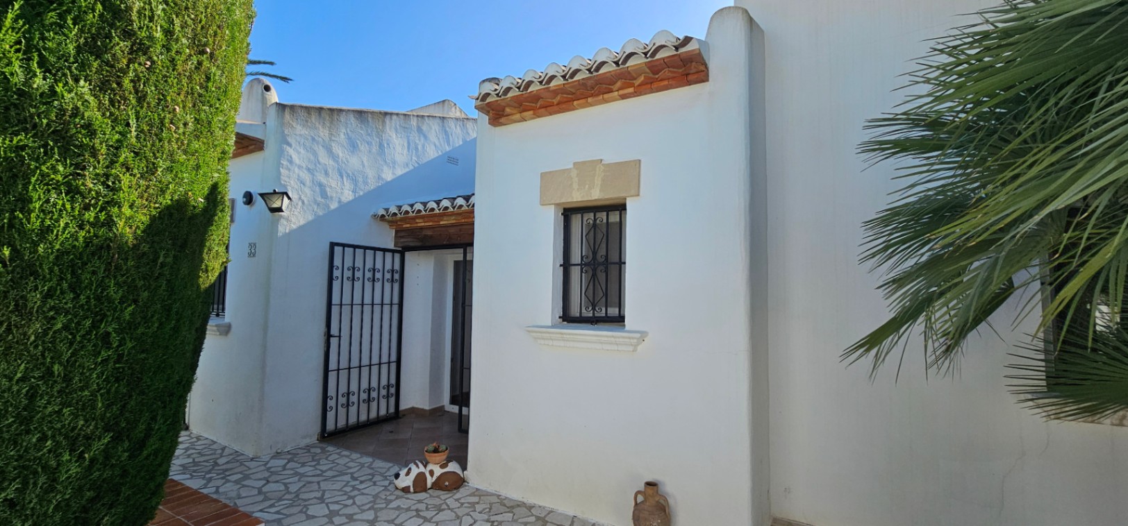 Exterior de villa blanca con tejas de terracota, ventana con rejas de hierro forjado y dintel de piedra. Entrada principal con portón de hierro y seto verde.