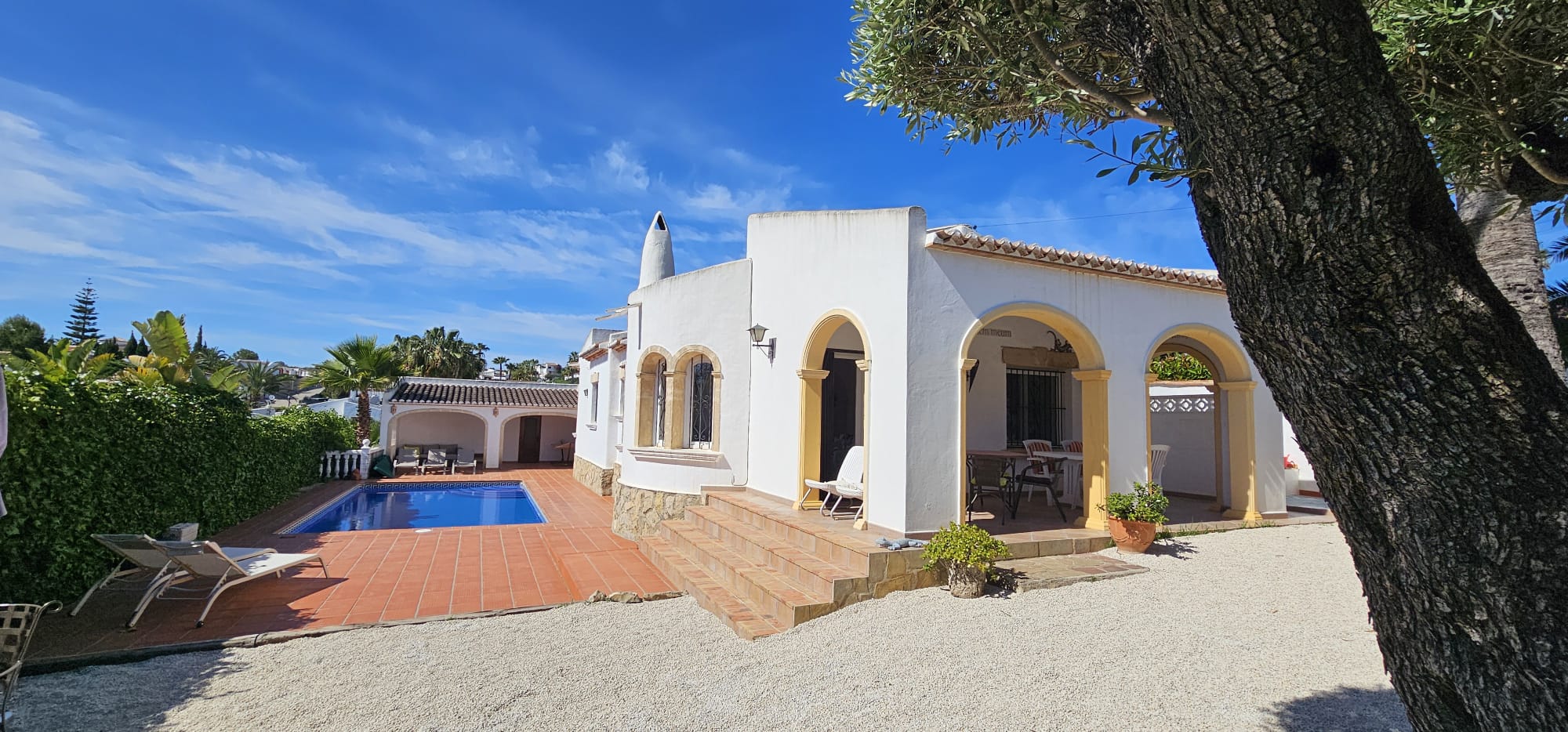 Exterior de villa mediterránea con fachada blanca y arcos amarillos. Destaca la piscina con solárium de terracota y una terraza cubierta para comedor al aire libre.
