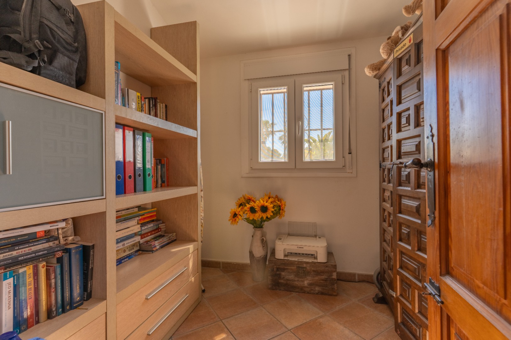 Habitación con estantería de madera clara, ventana de PVC de doble cristal con rejas y vista a palmeras. Puerta de madera tallada y suelo de baldosas.