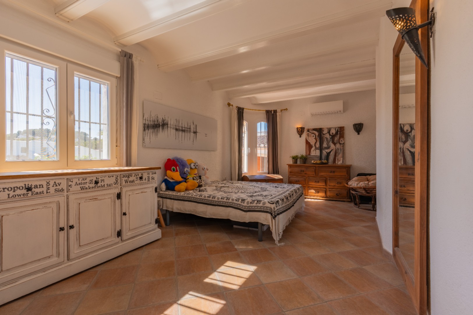 Luminosa estancia con suelo de terracota y vigas blancas. Destaca un mueble blanco envejecido junto a ventanas de PVC con rejas, inundada de luz natural.