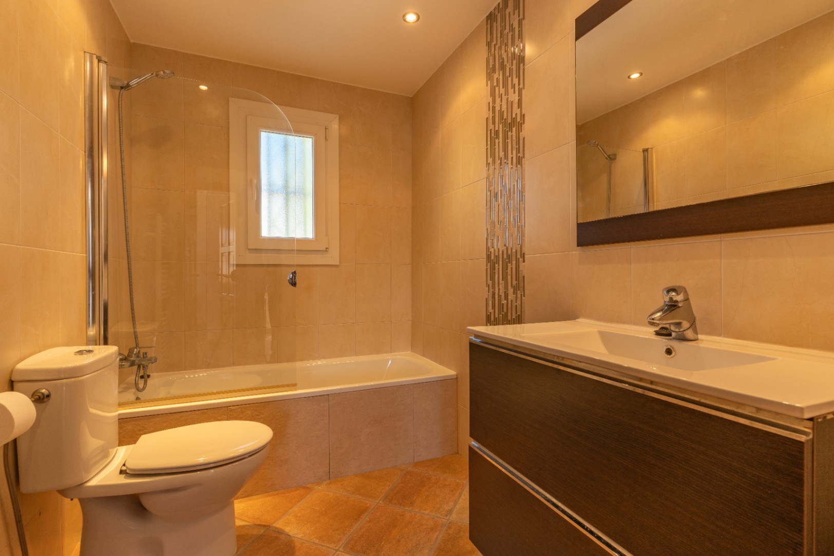 Luminoso baño con bañera y mampara de cristal, mueble de lavabo efecto madera oscura con seno blanco, y azulejos beige con franja decorativa de mosaico.