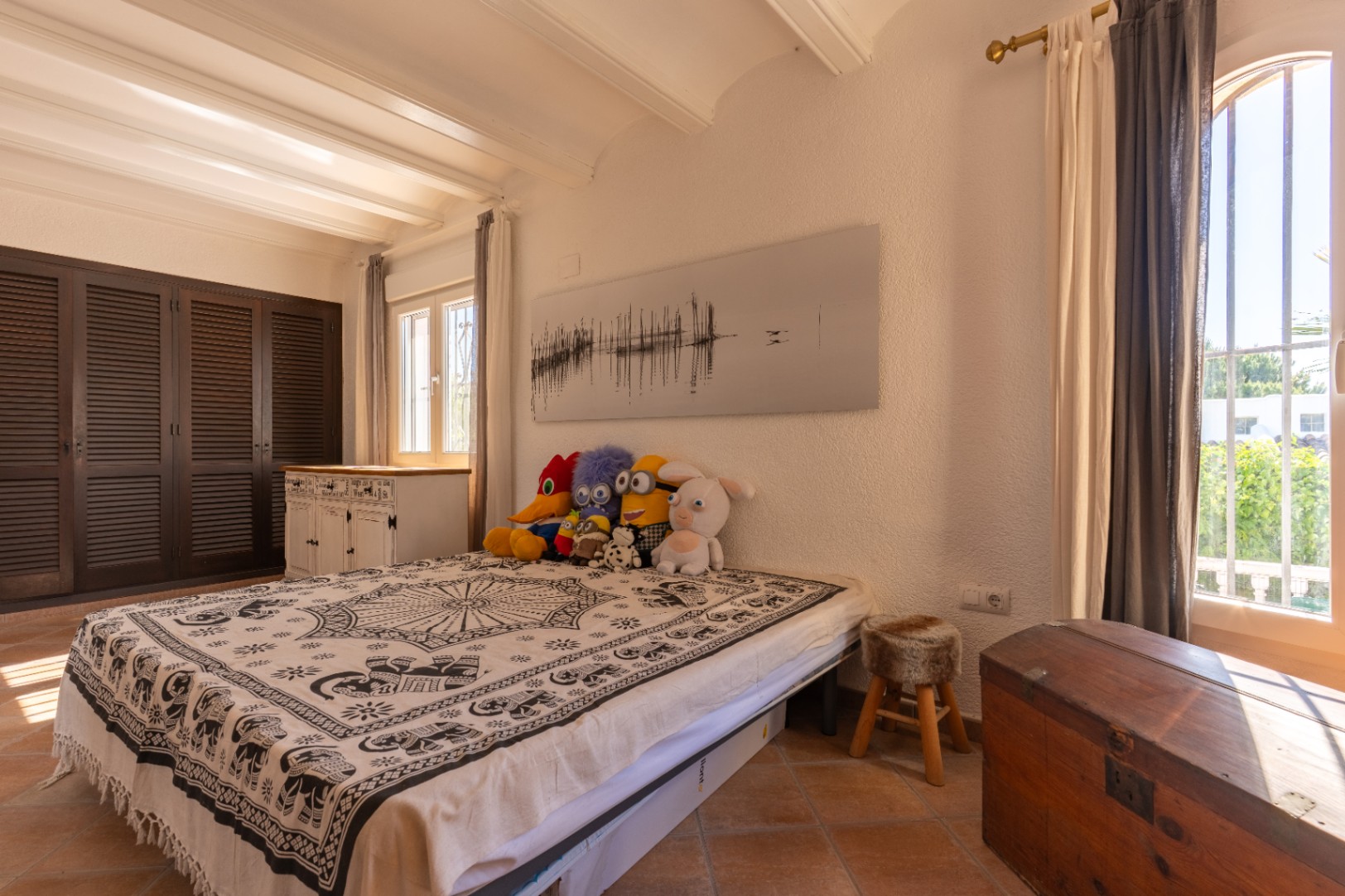Luminoso dormitorio con suelos de terracota, paredes blancas texturizadas y vigas de techo vistas. Dispone de armarios de lamas oscuras, cama con colcha estampada y luz natural de ventanas de PVC de doble cristal.