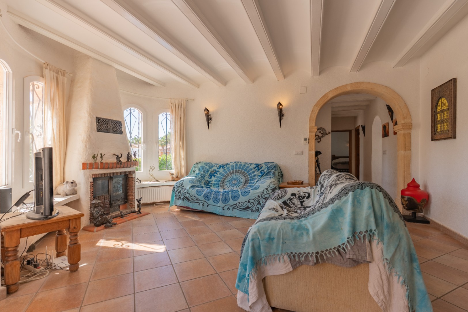 Luminoso salón con una chimenea tradicional de ladrillo, ventanas arqueadas con rejas y techo con vigas vistas. El suelo es de baldosas de terracota, y un arco de piedra decorativo conecta con el interior.