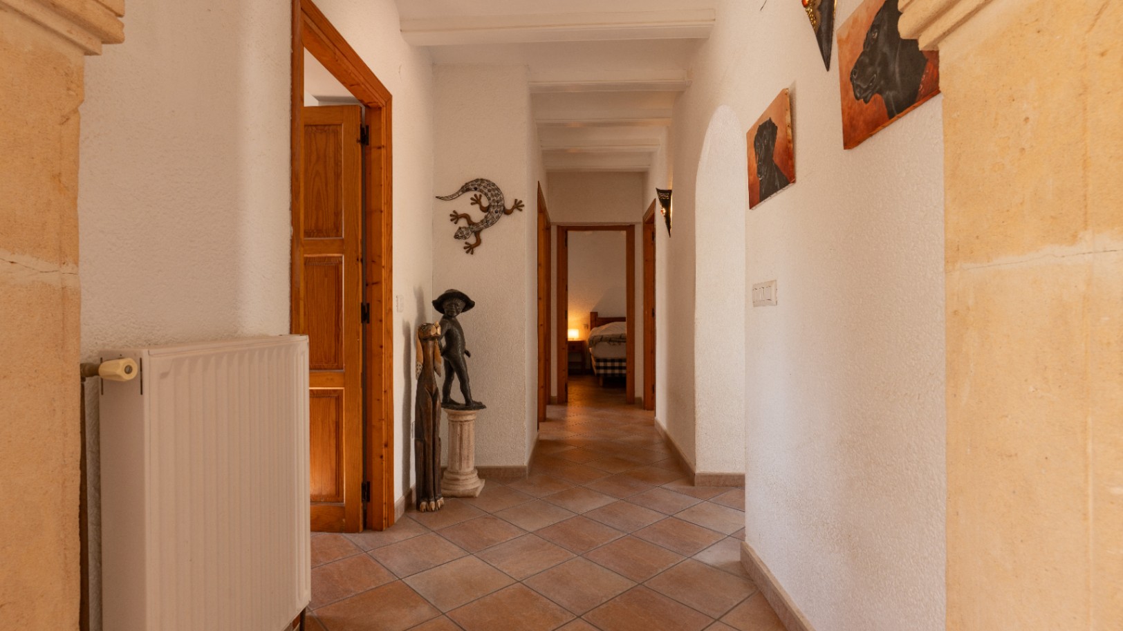 Pasillo luminoso con suelo de baldosas de terracota, paredes blancas texturizadas y vigas de madera vistas. Incluye radiador, elementos decorativos y vista a un dormitorio.