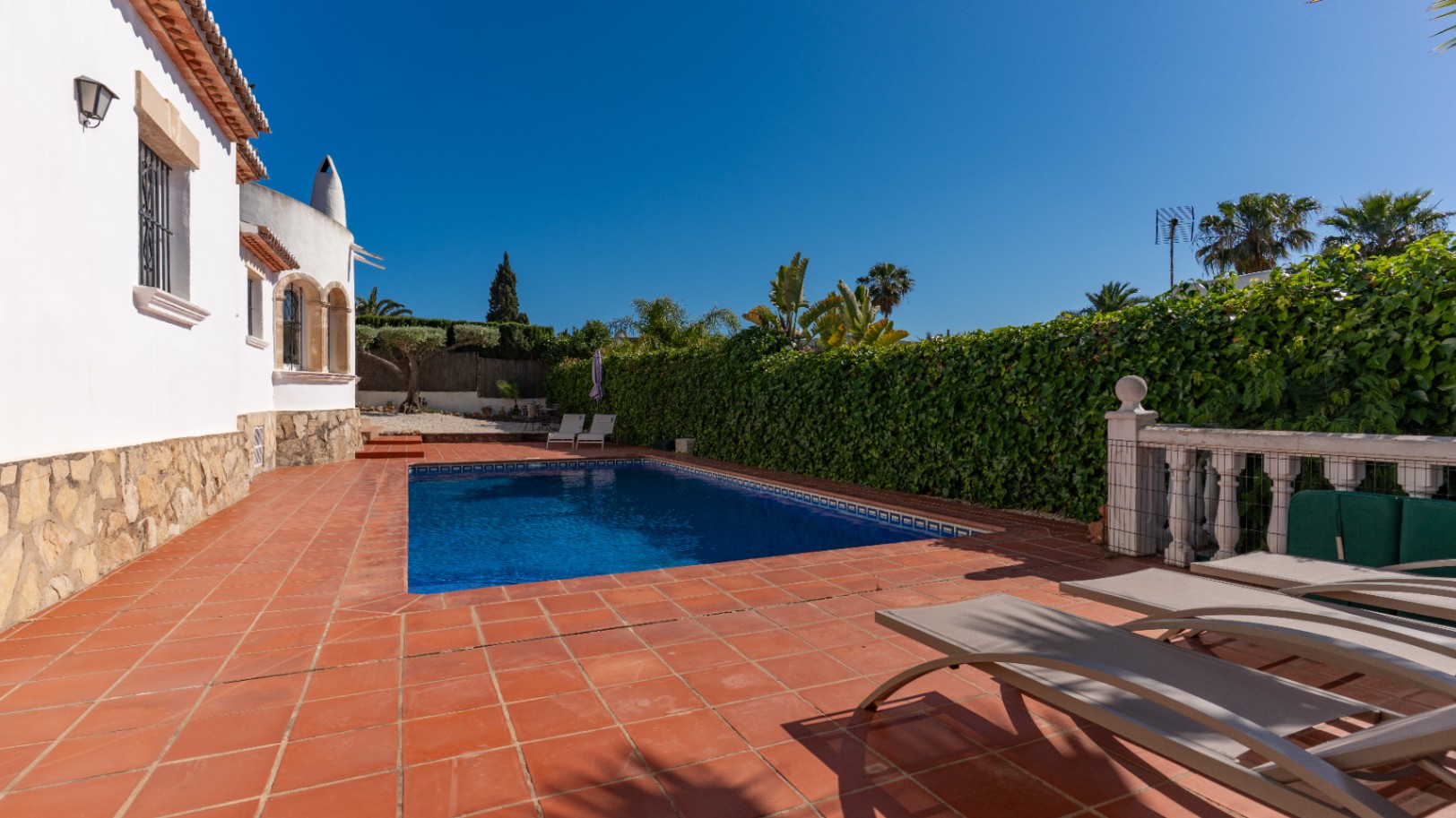 Piscina rectangular de azulejos azules en patio de terracota con tumbonas. Un seto alto y la villa blanca con zócalo de piedra enmarcan este espacio exterior privado.