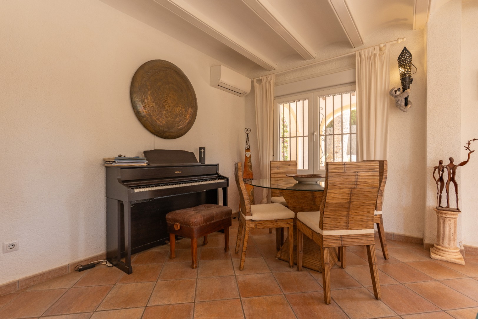 Rincón del comedor con suelo de baldosas de terracota, piano digital y mesa de cristal con sillas de bambú. La ventana de doble cristal con rejas ofrece luz natural.