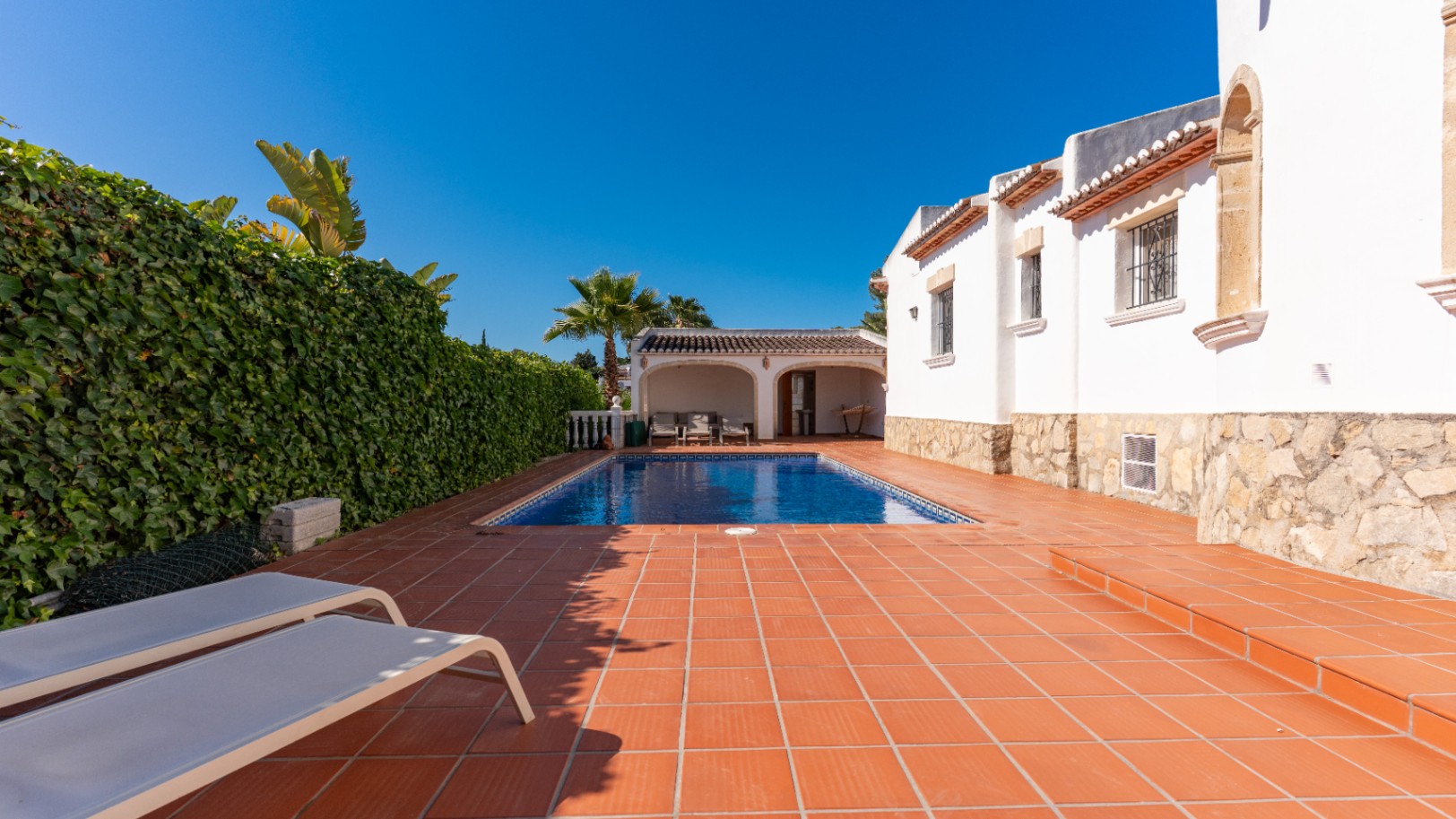 Soleada zona de piscina privada con piscina rectangular, terraza de baldosas de barro y dos tumbonas. Un seto verde denso ofrece privacidad junto a la villa blanca.