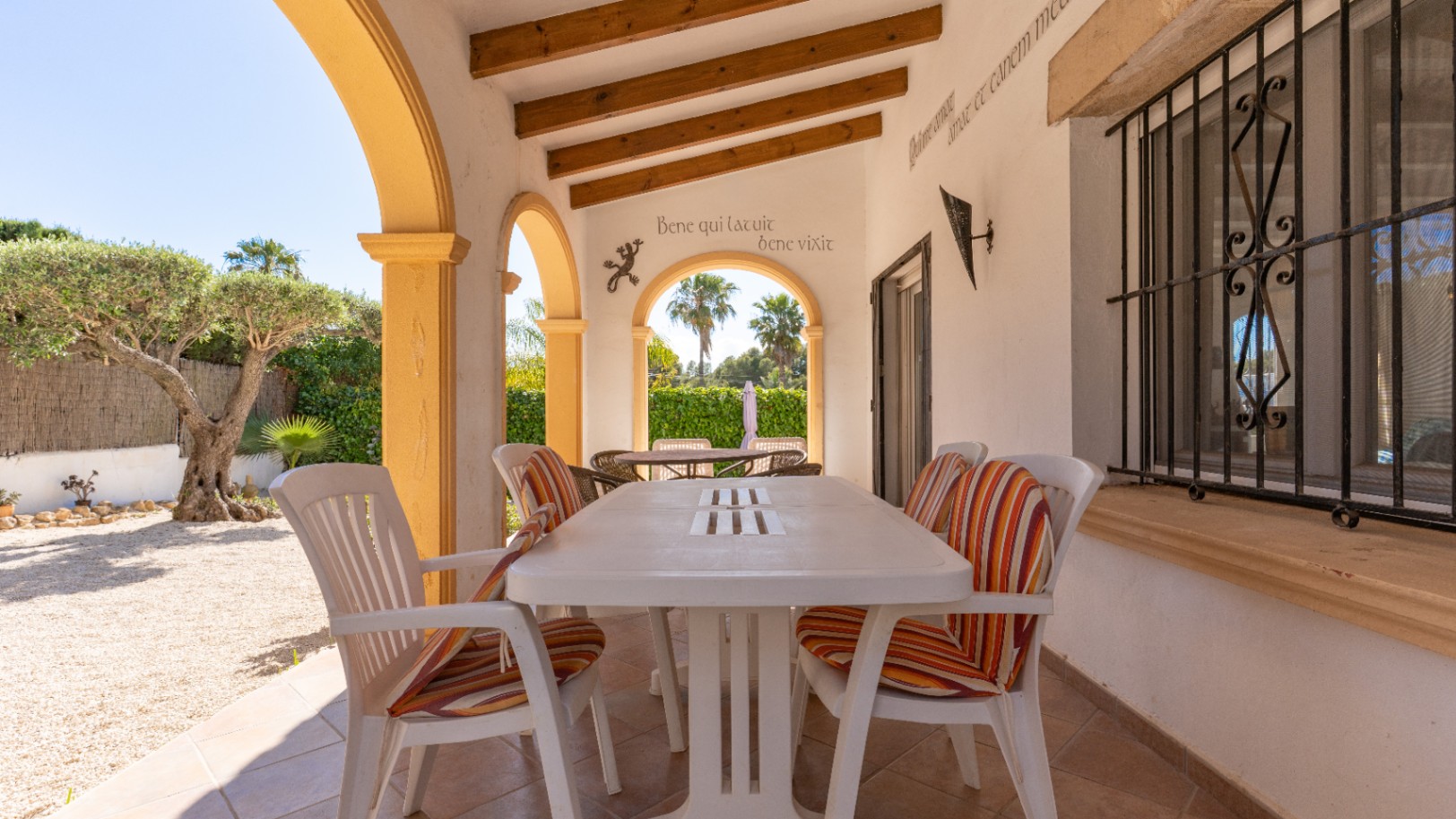 Terraza cubierta con suelo de baldosas, mesa de comedor blanca y sillas con cojines. Arcos amarillos y vigas de madera en el techo, con vista a un jardín de grava con olivo.