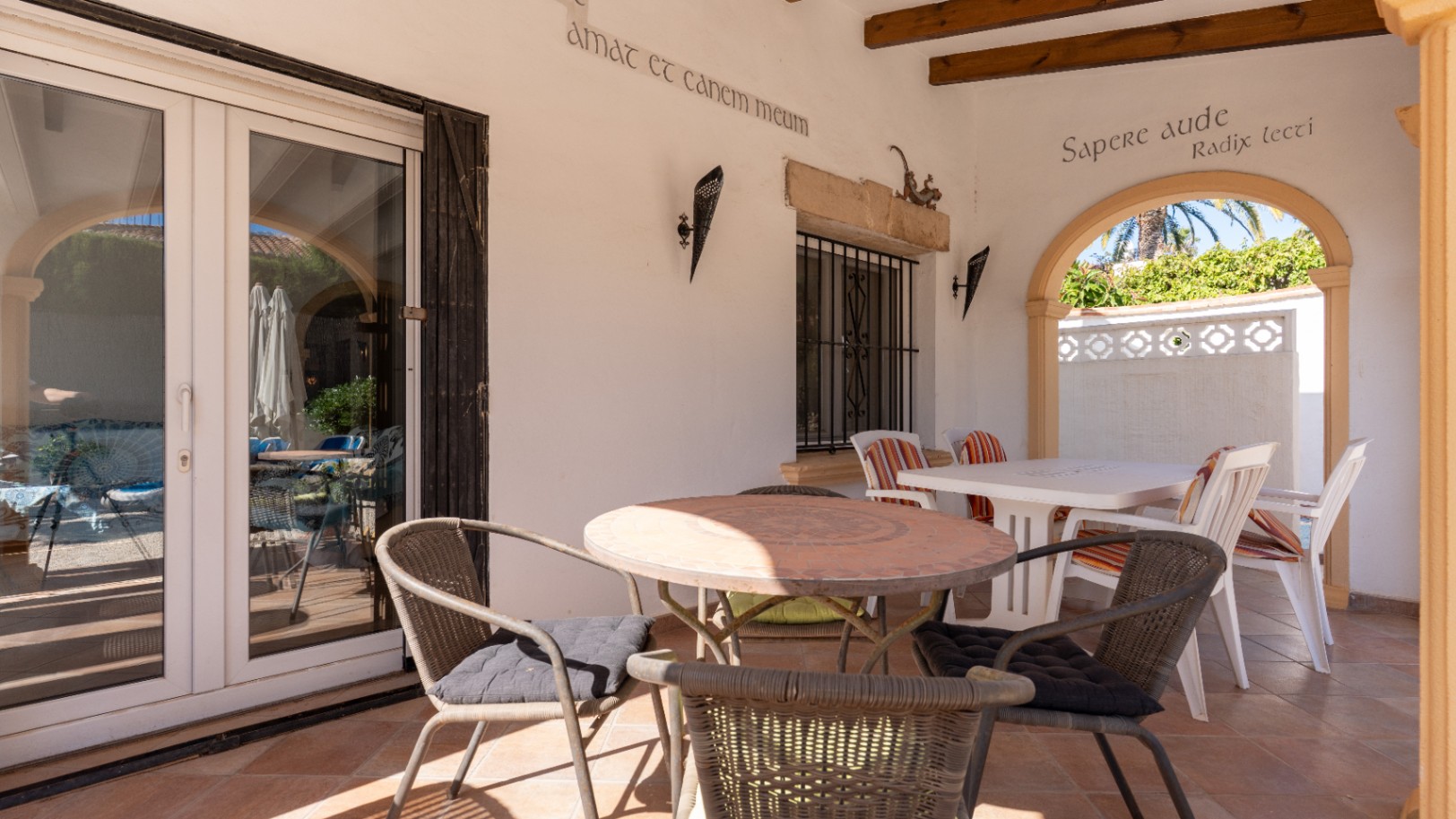 Terraza cubierta con suelos de terracota, mesa redonda de mosaico y puertas de PVC blanco. Un arco revela vegetación exterior y palmeras.