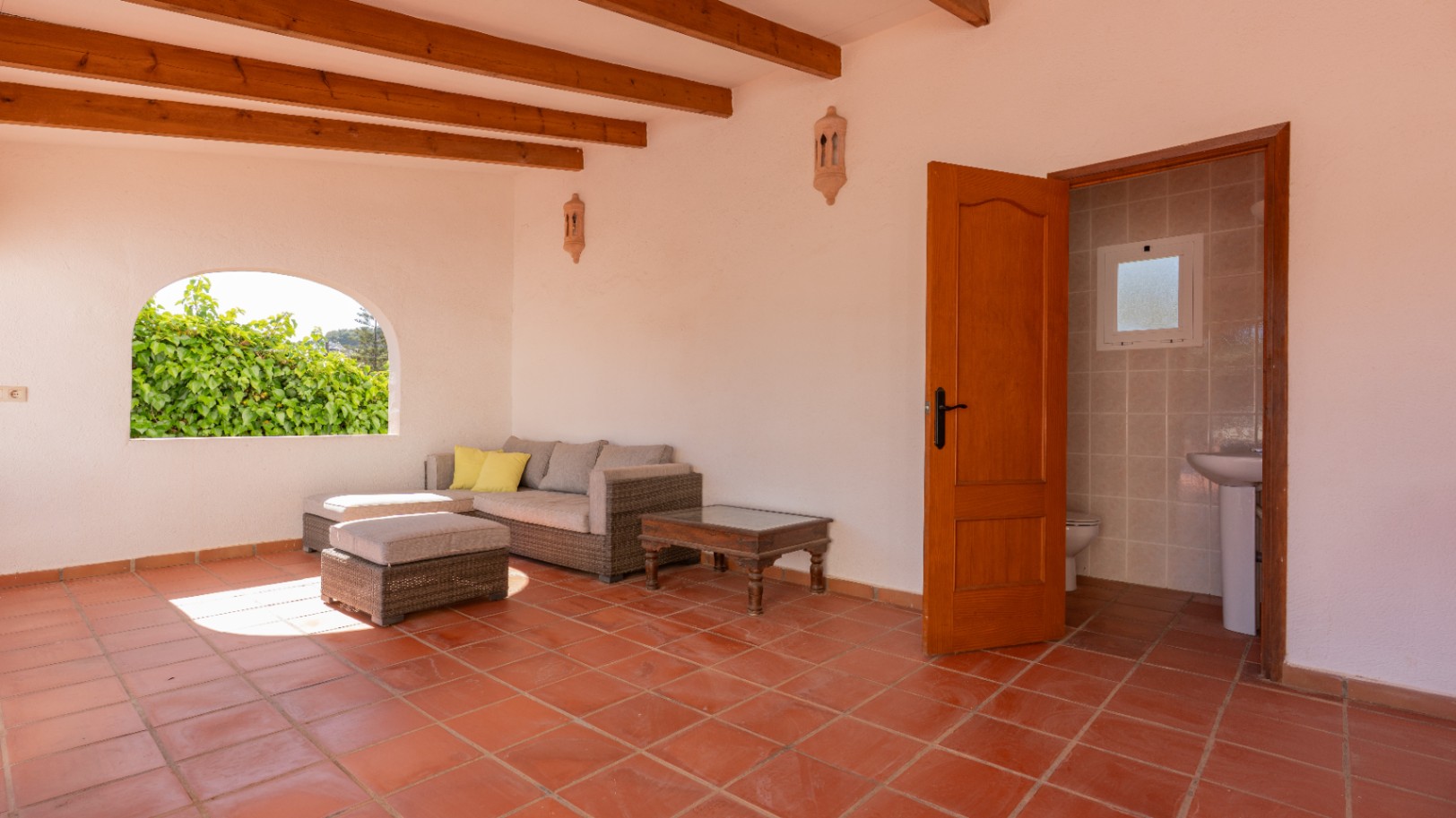 Terraza cubierta con suelos de terracota, vigas de madera y un sofá de ratán. Incluye un acceso a un aseo y una ventana arqueada con vistas a la vegetación.