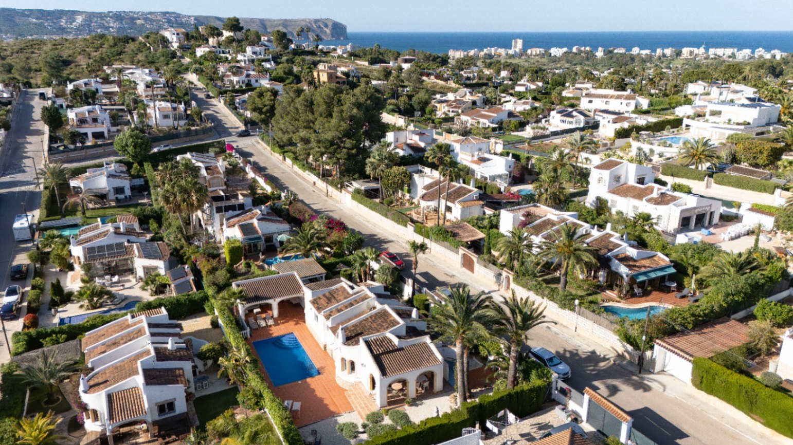 Vista aérea de la tranquila zona residencial de Jávea, mostrando villas blancas con piscinas privadas, exuberantes jardines mediterráneos y el mar azul a lo lejos.