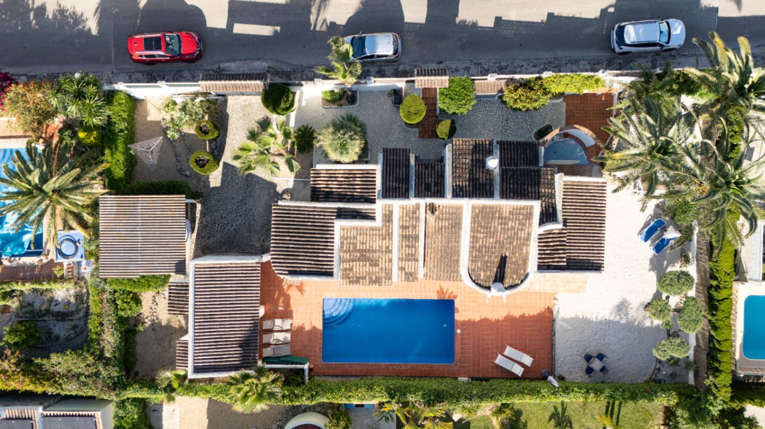 Vista aérea de la villa privada con piscina rectangular azul, terraza solárium de baldosas de terracota y amplio jardín con árboles mediterráneos y entrada pavimentada.