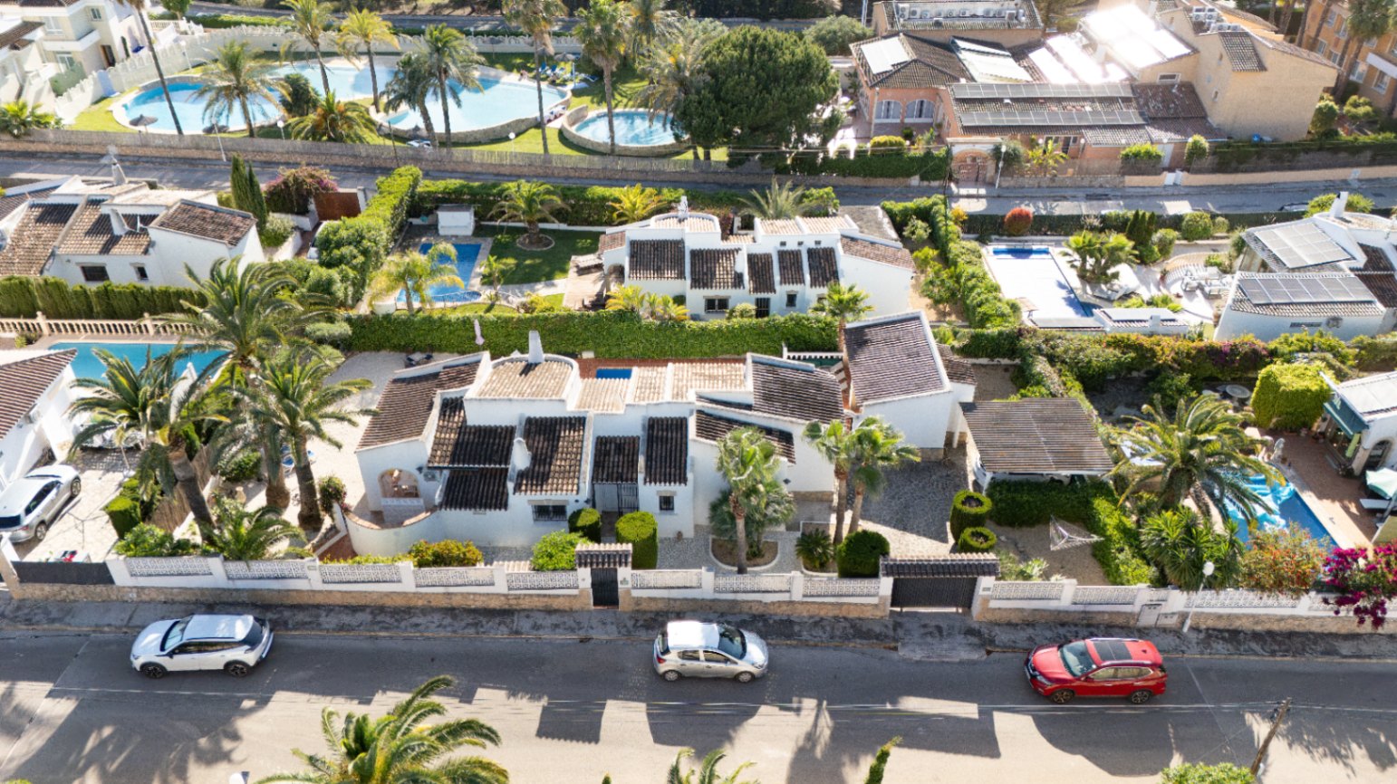 Vista aérea de la villa privada en Jávea, mostrando su fachada blanca, tejados oscuros, una gran piscina y un exuberante jardín mediterráneo con palmeras y entrada privada.