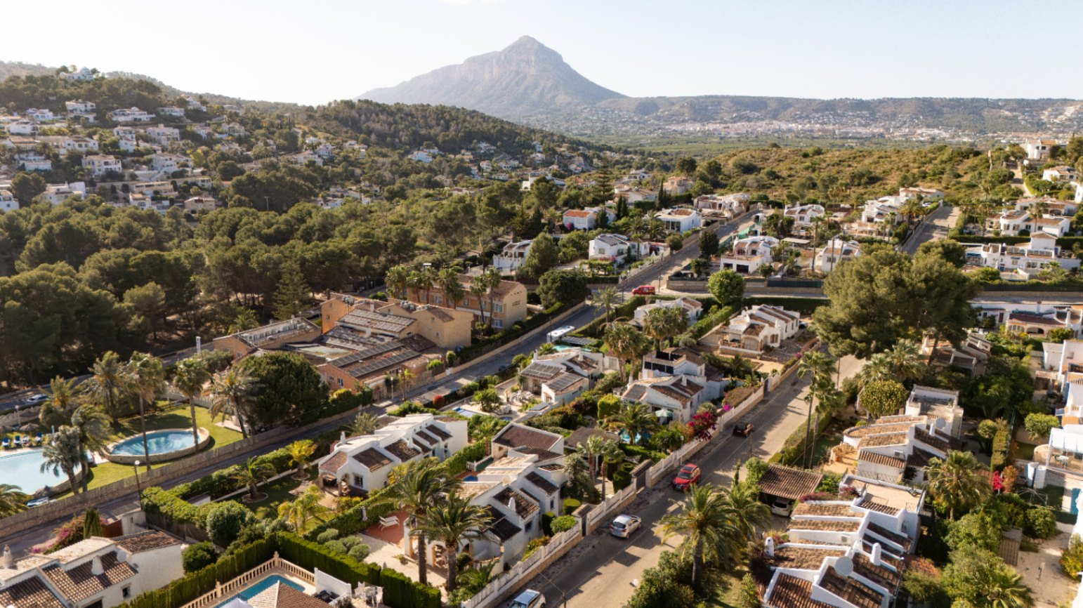 Vista aérea de una tranquila zona residencial, mostrando numerosas villas blancas con tejados de terracota, enclavadas entre abundante vegetación mediterránea y calles serpenteantes, con una montaña destacada al fondo.