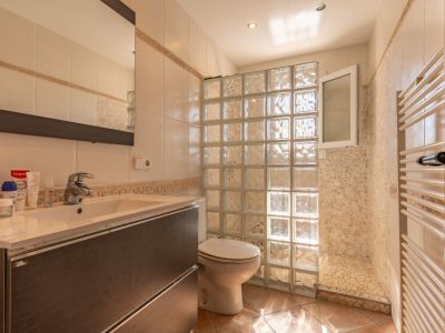 Baño funcional con mueble de lavabo oscuro, mampara de bloques de vidrio para la ducha y zona de ducha con azulejos de mosaico. Incluye toallero calefactado y suelo de terracota.