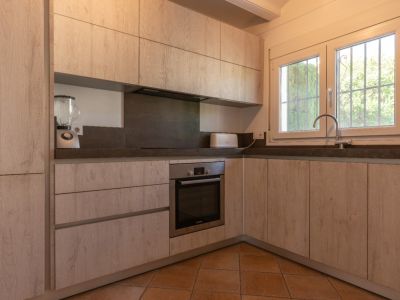 Cocina renovada en L con armarios efecto madera clara, encimeras grises oscuras y horno integrado. Ventana de PVC blanco con rejas aporta luz natural.