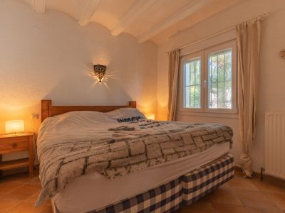 Dormitorio con cama de madera y base a cuadros, suelos de terracota y ventana de PVC con doble cristal. Destaca el aplique de pared con luz decorativa y el techo con vigas vistas.