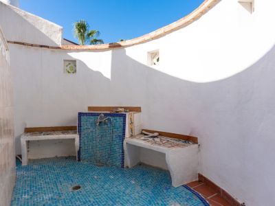 Ducha exterior con paredes blancas enlucidas y suelo de mosaico azul. Incluye bancos integrados con azulejos decorativos y una pared curva bajo el cielo azul.