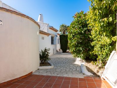 Entrada a la propiedad con un camino de piedra irregular, flanqueado por una pared curva blanca con celosía y exuberante vegetación mediterránea. Suelos de terracota en primer plano.