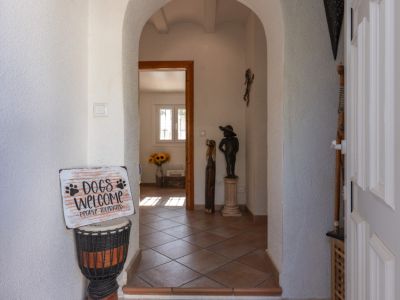 Entrada principal con arcos enlucidos en blanco y suelo de baldosas de terracota. La puerta de entrada panelada se ve a la derecha, y al fondo se divisa una estancia con luz natural y girasoles.