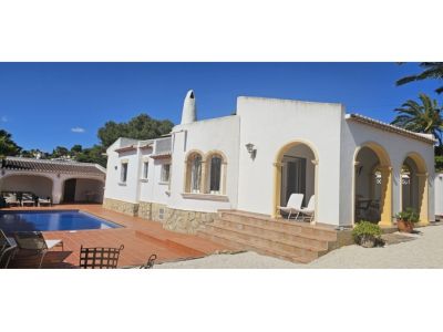 Exterior de villa blanca con arquitectura mediterránea, destacando una terraza cubierta con arcos ocres y escaleras de terracota. Piscina azul y zona de aparcamiento lateral.