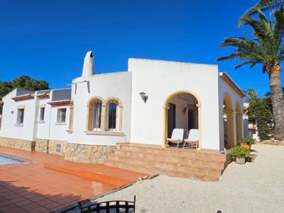 Exterior de villa blanca con base de piedra, escalones de terracota y porche arqueado. Piscina con terraza de gres y jardín mediterráneo con palmeras bajo cielo azul.