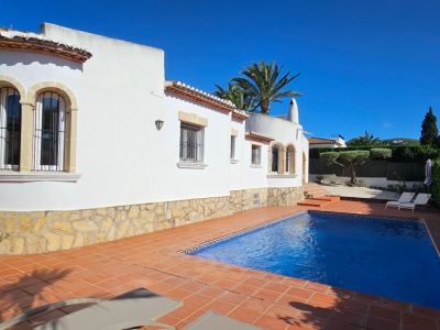 Exterior de villa blanca con base de piedra y ventanas arqueadas con rejas. Piscina rectangular rodeada de terraza de barro cocido, palmeras y vegetación bajo un cielo azul.