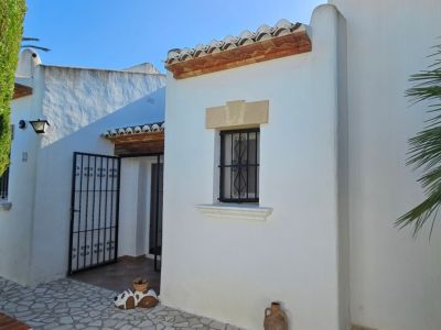 Exterior de villa blanca con tejas de terracota, ventana con rejas de hierro forjado y dintel de piedra. Entrada principal con portón de hierro y seto verde.