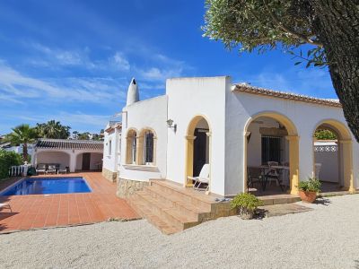 Exterior de villa mediterránea con fachada blanca y arcos amarillos. Destaca la piscina con solárium de terracota y una terraza cubierta para comedor al aire libre.
