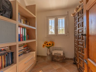 Habitación con estantería de madera clara, ventana de PVC de doble cristal con rejas y vista a palmeras. Puerta de madera tallada y suelo de baldosas.