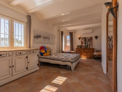 Luminosa estancia con suelo de terracota y vigas blancas. Destaca un mueble blanco envejecido junto a ventanas de PVC con rejas, inundada de luz natural.