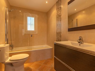 Luminoso baño con bañera y mampara de cristal, mueble de lavabo efecto madera oscura con seno blanco, y azulejos beige con franja decorativa de mosaico.