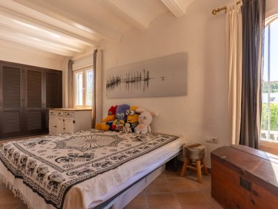Luminoso dormitorio con suelos de terracota, paredes blancas texturizadas y vigas de techo vistas. Dispone de armarios de lamas oscuras, cama con colcha estampada y luz natural de ventanas de PVC de doble cristal.