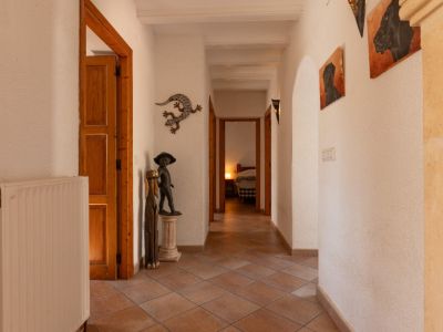 Pasillo luminoso con suelo de baldosas de terracota, paredes blancas texturizadas y vigas de madera vistas. Incluye radiador, elementos decorativos y vista a un dormitorio.