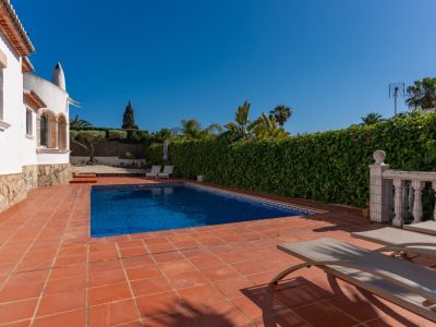 Piscina rectangular de azulejos azules en patio de terracota con tumbonas. Un seto alto y la villa blanca con zócalo de piedra enmarcan este espacio exterior privado.