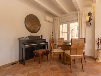 Rincón del comedor con suelo de baldosas de terracota, piano digital y mesa de cristal con sillas de bambú. La ventana de doble cristal con rejas ofrece luz natural.