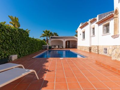 Soleada zona de piscina privada con piscina rectangular, terraza de baldosas de barro y dos tumbonas. Un seto verde denso ofrece privacidad junto a la villa blanca.