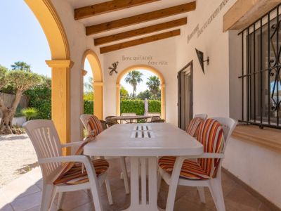 Terraza cubierta con suelo de baldosas, mesa de comedor blanca y sillas con cojines. Arcos amarillos y vigas de madera en el techo, con vista a un jardín de grava con olivo.